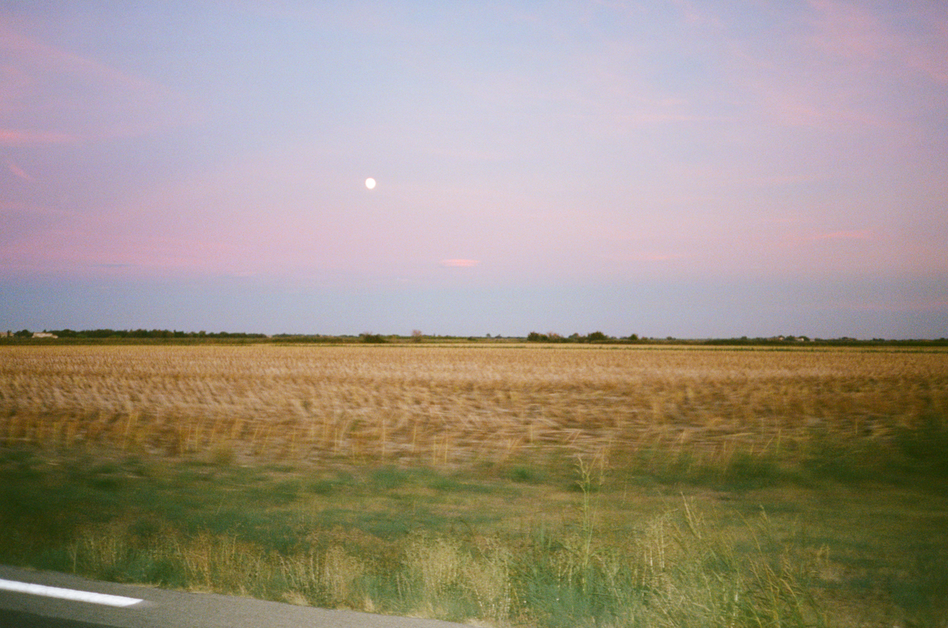 Photographie argentique couleur prise sur la route en voiture du côté de la route. On y voit un champ et le ciel es orangé. On aperçoit également la lune.