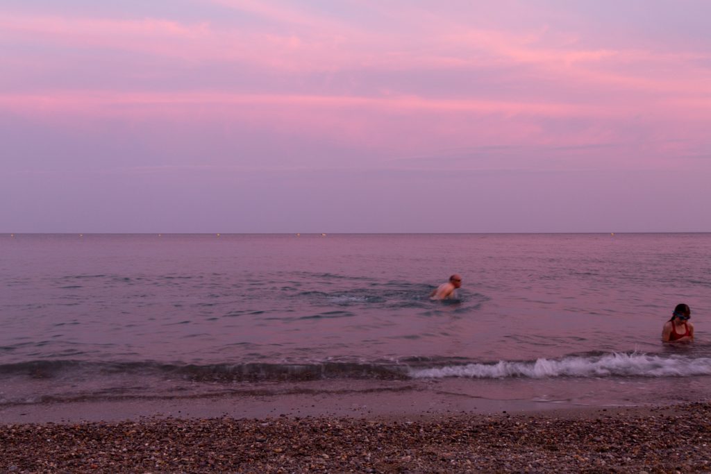 Photographie au couché de soleil de la mer Méditerrannée. On remarque une petite fille dans l'eau à droite de l'image et un monsieur flou qui semble faire la nage du papillon plus au centre de l'image.