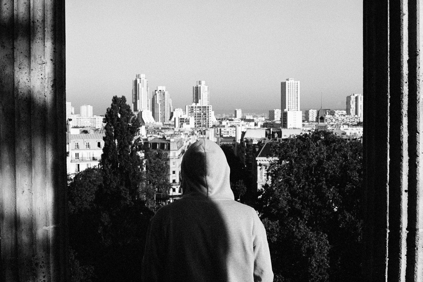 Photographie argentique noir et blanc avec une personne portant une capuche vue de dos regardant le paysage parisien depuis le point culminant du parc des Buttes Chaumonds à Paris. Les bords verticaux de l'image sont délimités par les pillones du kiosque. Une ombre traverse le dos du personnage de manière verticale.